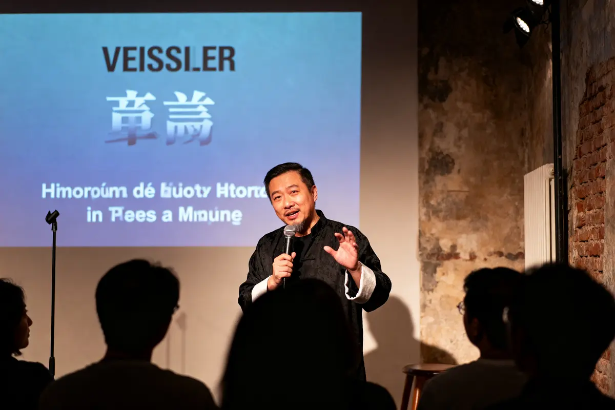 Homme parlant au micro sur scène, écran avec texte en arrière-plan, audience visible au premier plan.