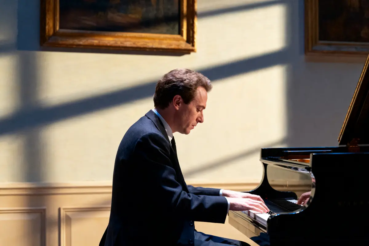 Homme jouant du piano dans une pièce éclairée par la lumière du jour, portant un costume sombre.